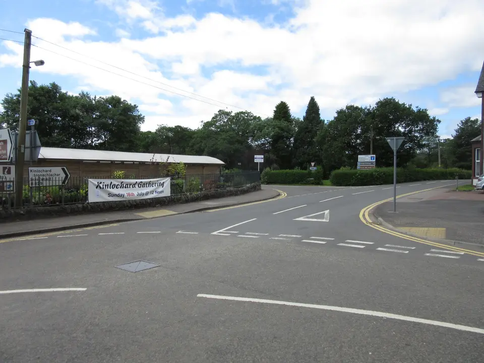 A road junction in Aberfoyle with a village sign, trees and a low building behind