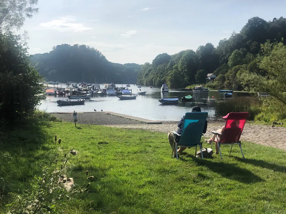 Balmaha harbour on a sunny day, with moored boats, a grassy shore and wooded hills behind