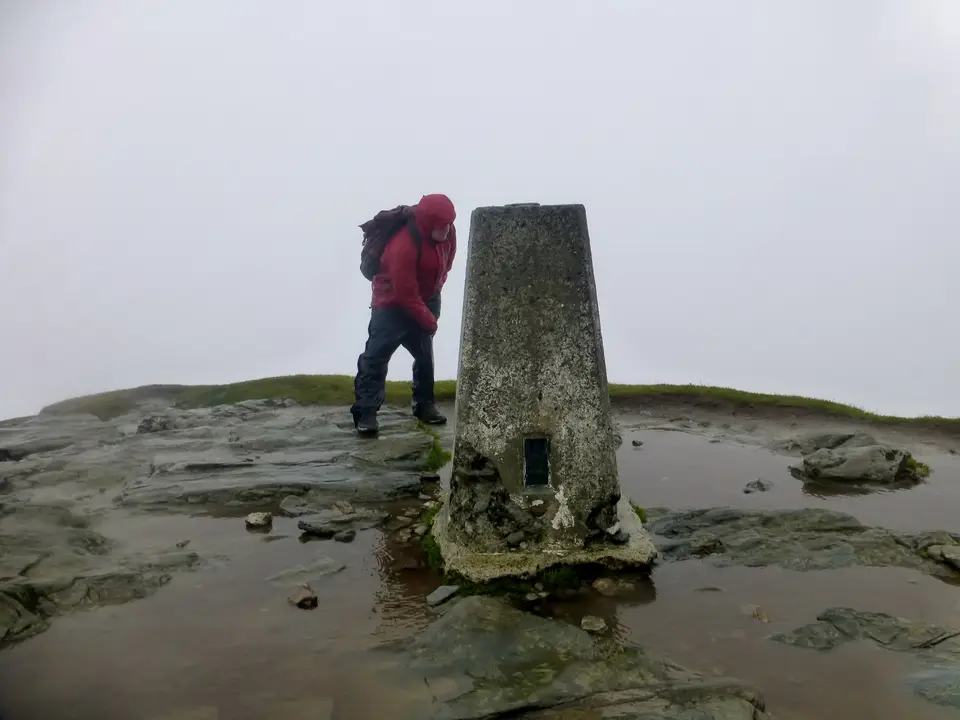 A walker in waterproofs beside the trig point on the summit of Ben Lomond in low cloud