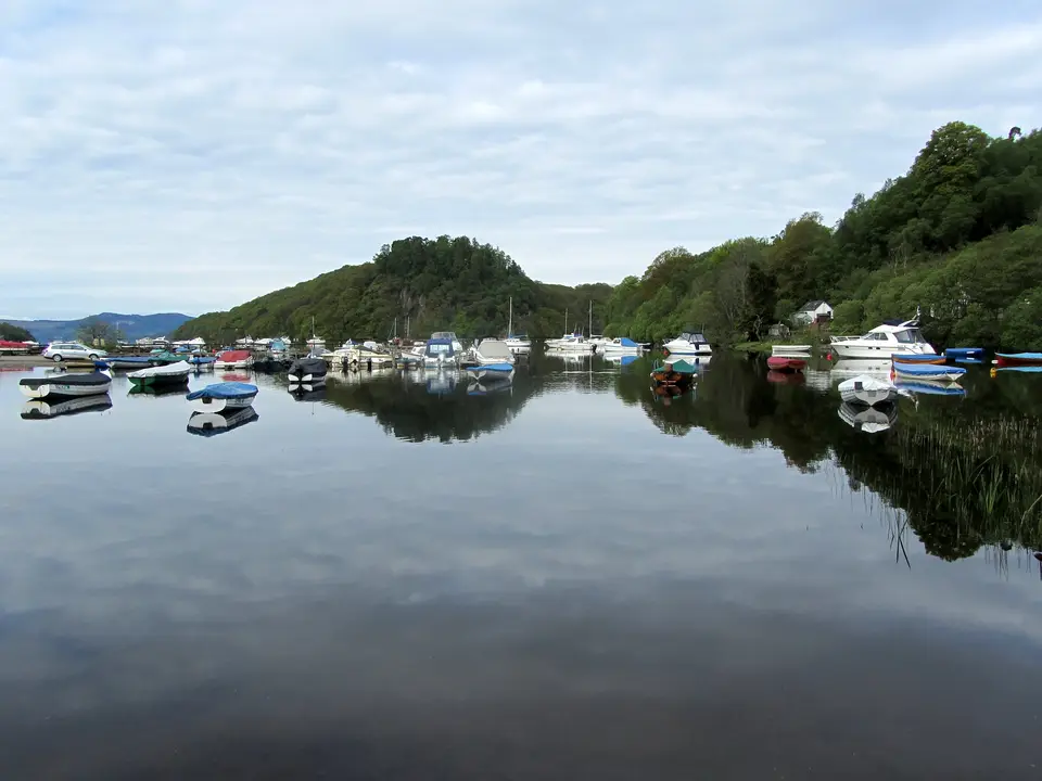 Boats moored in Balmaha Bay on a still, cloudy day, with a wooded headland behind