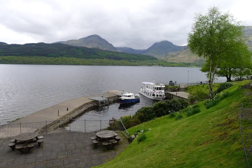 Boats moored at the Inversnaid harbour jetty with a wooded hill and mountains behind