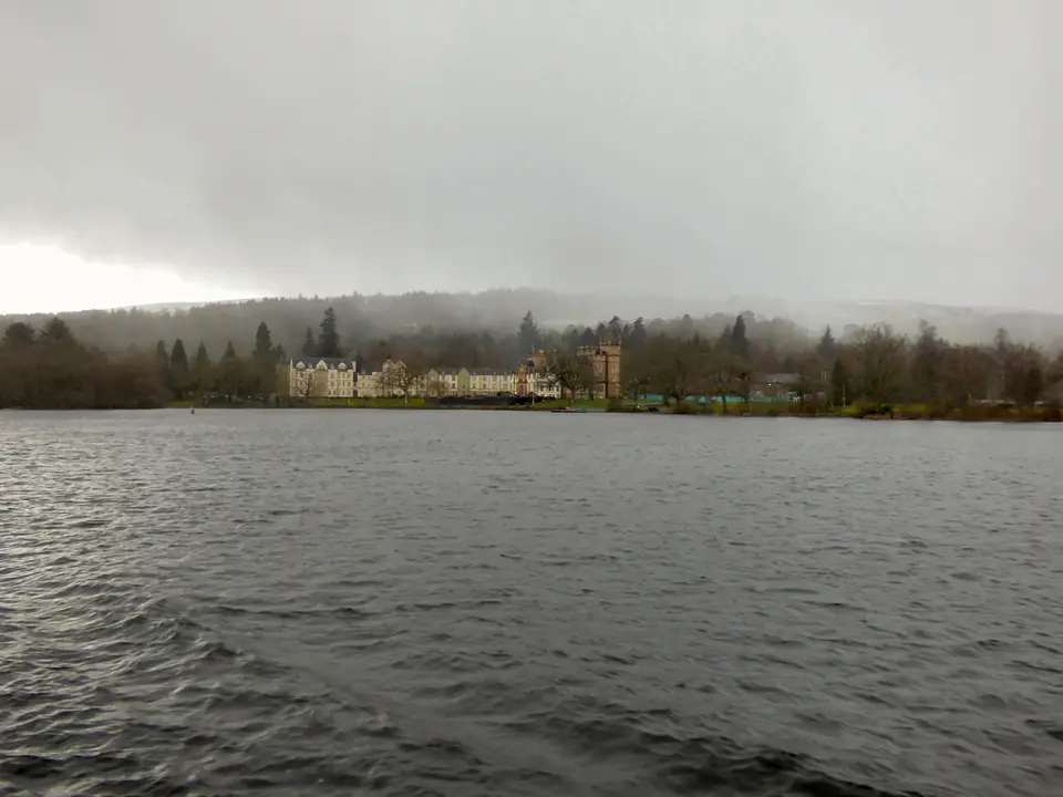 Cameron House hotel seen across the grey water of Loch Lomond, with trees and low cloud behind it