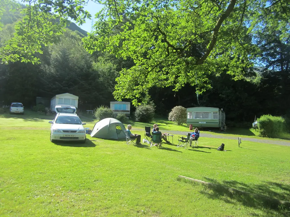 Campers sitting in chairs beside a tent and caravans on a grass campsite below wooded hills