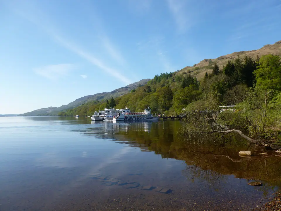 Cruise boats moored at a wooded Loch Lomond shore under a blue sky