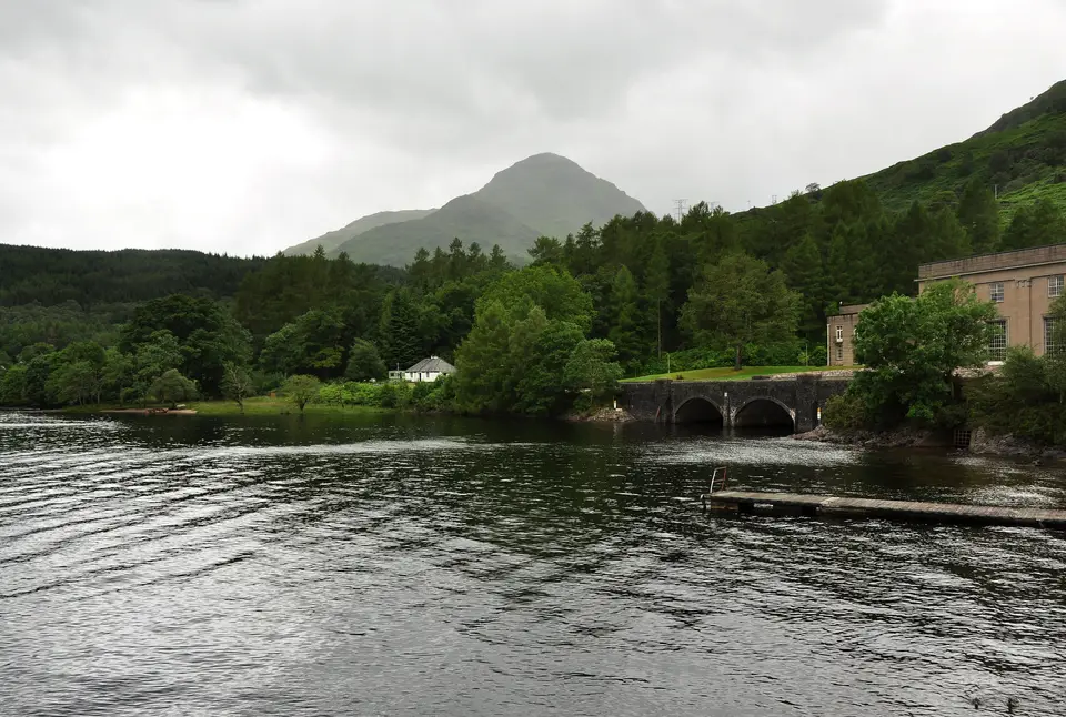 Wooded Inveruglas shore with the arched bridge, a power station building and a pointed hill behind