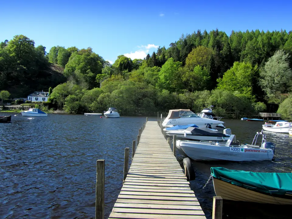 A wooden jetty with small boats moored alongside and wooded hills across the water