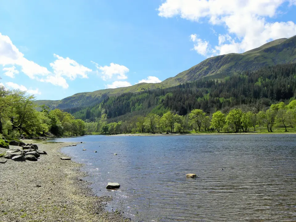 Loch Lubnaig on a bright spring day with a stony shore and forested hills rising behind