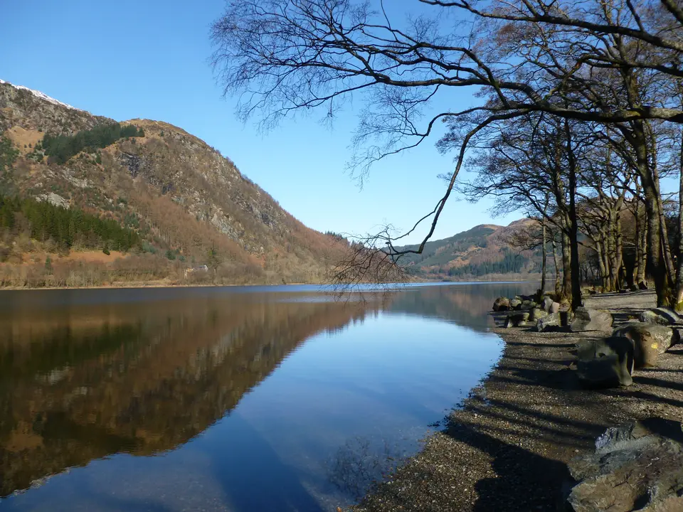 Still water on Loch Lubnaig reflecting a steep wooded hillside, with bare trees on a pebbly shore