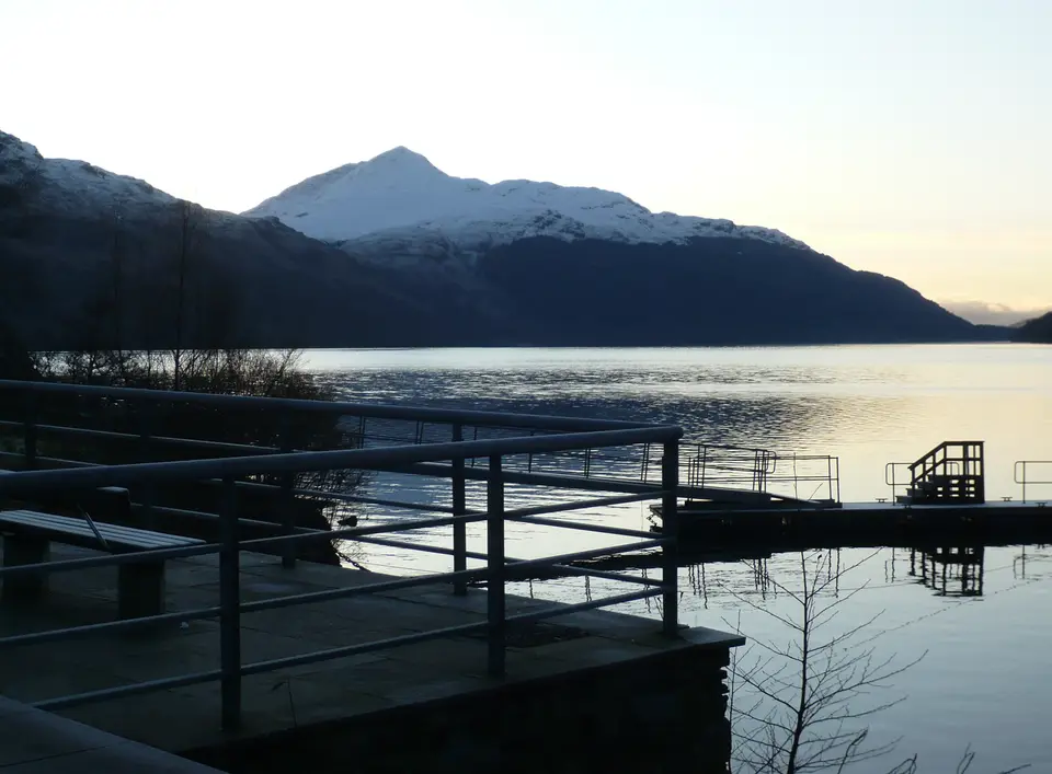 Ben Lomond with snow on its top seen across calm water from a railed lochside deck at dusk