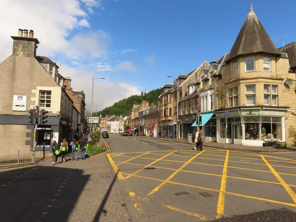 Main Street in Callander on a sunny day, with a corner building with a pointed turret and a wooded hill behind