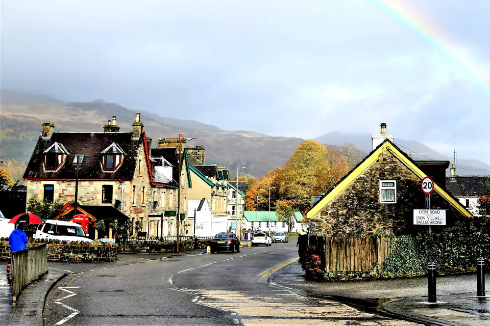 Stone houses and shops on Killin Main Street under a rainbow with hills rising behind the village