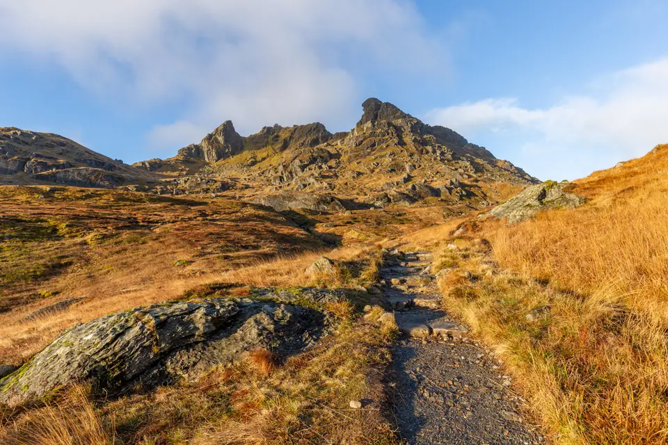 A stony path climbing towards the three rocky peaks of the Cobbler in low autumn light