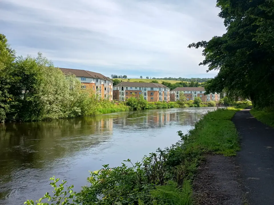 The River Leven flowing beside a tarmac path and flats in Alexandria, with trees along the far bank
