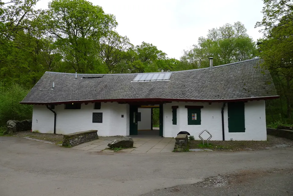 The white-walled public toilet block beside the tarmac of Rowardennan car park under trees