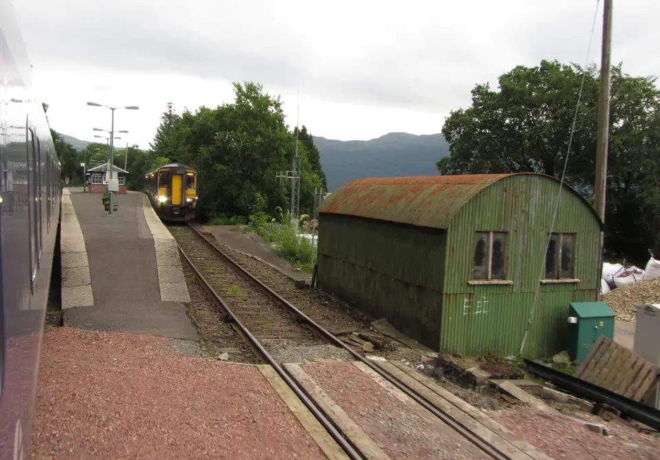 A train waiting at Arrochar and Tarbet station beside a green corrugated shed, with wooded hills behind
