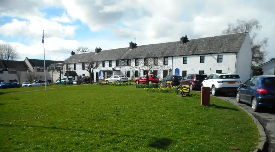 The long white Winnock building facing the green on The Square in Drymen, with parked cars in front